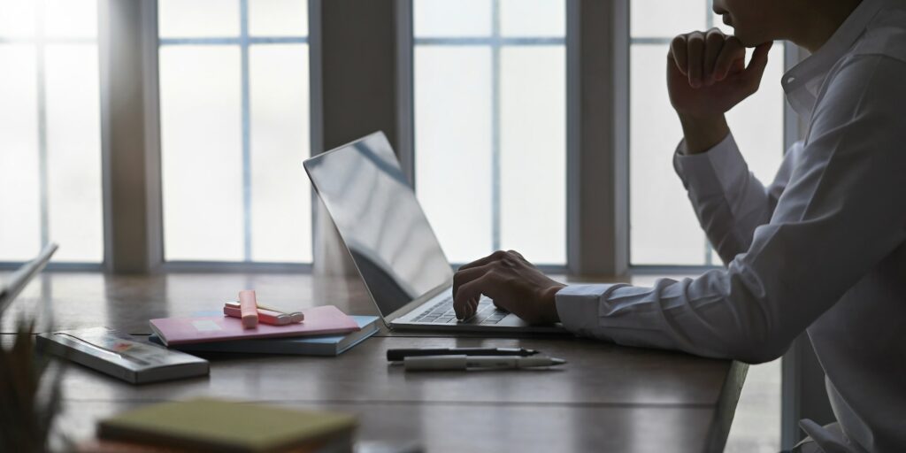 Man on white shirt keep his hand on chin while thinking and sitting in front a computer.
