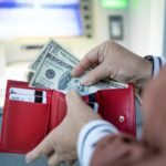 Woman hand puts many dollars in wallet near ATM outdoor. Modern technology, banking money